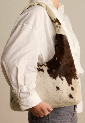 Bruin en witte, gestipte handtas van bont, met een zachte textuur en een gebogen ontwerp, onder de arm gedragen door een persoon in een wit shirt.