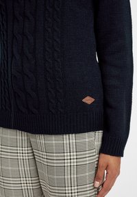 Maglione lavorato a maglia navy con motivi a treccia e polsini a costine. Include una patch in pelle marrone con il logo sul lato inferiore. Abbinato a pantaloni a quadri.