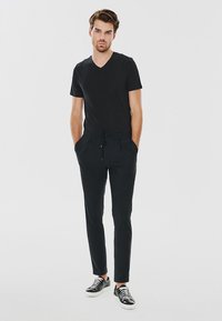 Czarny t-shirt z dekoltem w serek i dopasowane czarne spodnie z troczkami; model w czarno-białych sandałach, stojący z rękami w kieszeniach.