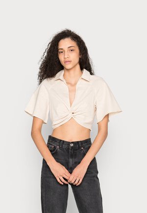 Camisa curta em bege claro com design de frente torcido e mangas curtas, combinada com jeans pretos de cintura alta. Tecido suave e texturizado.