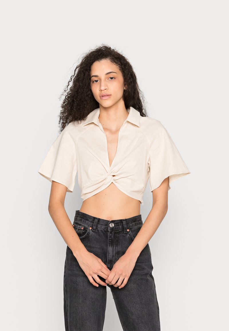Camisa curta em bege claro com design de frente torcido e mangas curtas, combinada com jeans pretos de cintura alta. Tecido suave e texturizado.