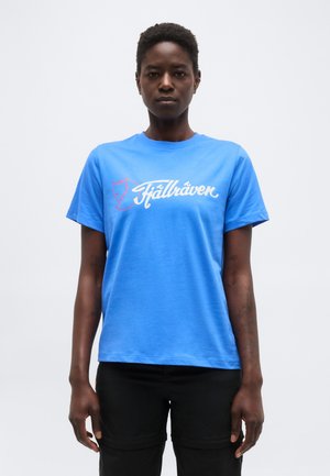 Person wearing a blue Fjällräven t-shirt and black pants, standing facing forward against a plain white background.