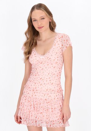 Młoda kobieta z długimi, falowanymi włosami, ubrana w różową sukienkę w kwiaty z krótkim rękawem i falbanami na spódnicy, stojąca na tle białego tła.