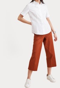 Weißes kurzärmliges Button-Up-Hemd, kombiniert mit rostfarbenen cropped Hosen. Das Model trägt weiße Sneakers. Glatte Texturen und moderner Schnitt.