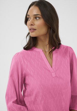 Žena s dlhými tmavými vlasmi, oblečená v textúrovanej jasne ružovej blúzke a jemnom striebornom náhrdelníku, pozerajúca sa doľava na jednoduchom pozadí.