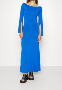 Blå, långärmad klänning med båturringning och sprund vid handlederna. Slät tyg, figurnära design, maxilängd. Svarta platåsandaler.