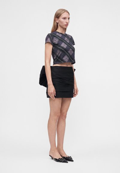 Femme aux longs cheveux blonds portant un top court à carreaux noir et gris, une mini-jupe noire avec détail superposé et des chaussures pointues noires avec des nœuds.