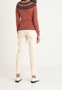 Un pull couleur rouille avec un empiècement noir et blanc à motifs, associé à un jean crème avec des ourlets effilochés et des chaussures noires à lacets.