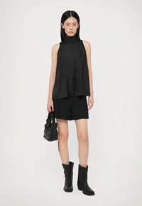 Vrouw staat en draagt een mouwloze zwarte turtleneck top, zwarte shorts, zwarte laarzen en houdt een kleine zwarte handtas met zilveren studs vast.