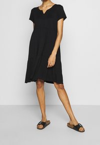 Robe noire à manches courtes avec un décolleté en V, un design à étages et une coupe décontractée. Complétée par des sandales noires à enfiler ornées de ferrures décoratives.