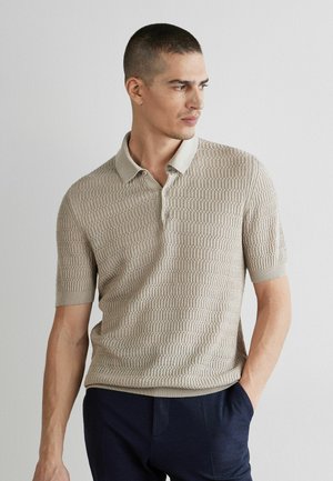 Mand iført beige kortærmet tekstureret strikket polo og mørke bukser, kigger mod højre med den ene hånd i lommen, foran en lys ensartet væg.