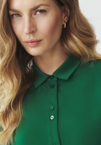 Green collared shirt with buttons, made of smooth fabric; model with wavy hair and earrings partially visible in a close-up view.