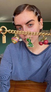 Bracelet en chaîne dorée orné de charms colorés : un ornement rayé, un dinosaure vert et des cerises rouges avec une finition texturée.