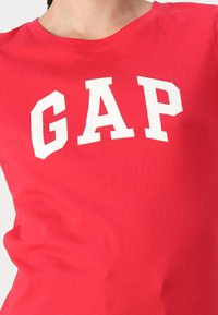 Raudona medvilninė marškinė su apvalia apykakle ir drąsiu baltu GAP logotipu spausdintu per krūtinę. Trumpomis rankovėmis ir minkšta tekstūra.