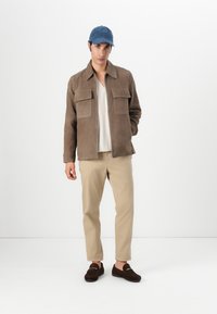 Brun suedjakke med to store brystlommer, hvid tekstureret skjorte under, beige bukser og marineblå kasket. Mørkebrune loafers fuldender looket.