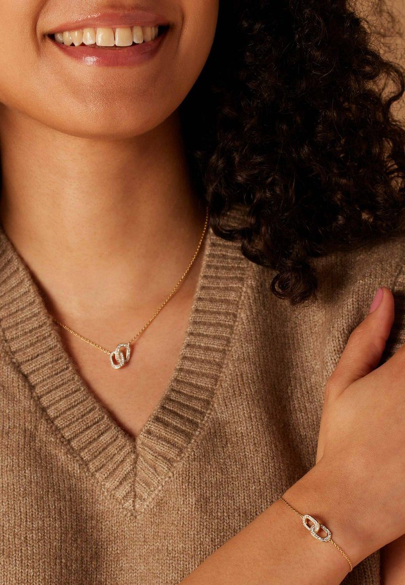 Gouden ketting en armband met een schakelsysteem, voorzien van een diamanten accent. Draagt in combinatie met een getextureerde bruine trui.