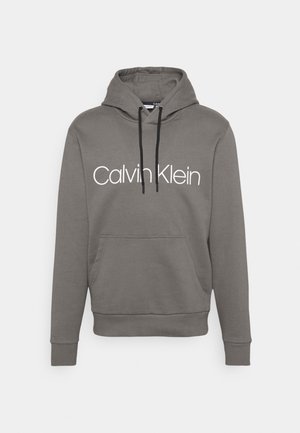 Szara bluza z kapturem z czarnymi sznurkami, przednią kieszenią i białym logo "Calvin Klein" na środku klatki piersiowej.