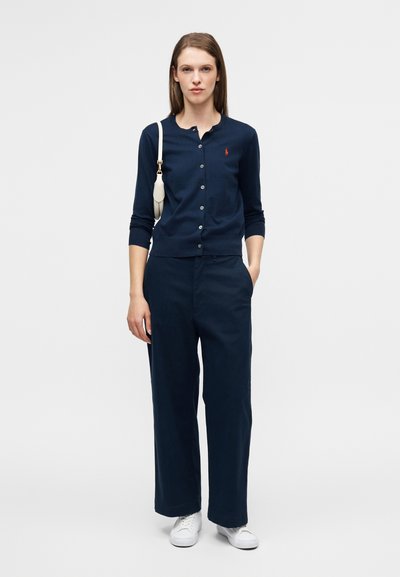 Καζάκα σε μπλε-ναυτικό χρώμα με κουμπιά και μακριά μανίκια, σετ με φαρδιά μπλε-ναυτικά παντελόνια. Το μοντέλο κρατά μια μικρή λευκή τσάντα και φορά λευκά αθλητικά παπούτσια.