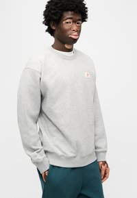 Γκρι φούτερ με στρογγυλή λαιμόκοψη, ριμπ μανσέτες και μικρό λογότυπο στο στήθος. Μαλακό, υφή υφασμα, στιλιζαρισμένο με τιρκουάζ παντελόνι.