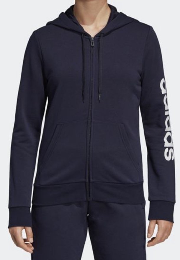 Haut de survêtement zippé bleu marine avec poches avant et logo Adidas imprimé verticalement sur la manche gauche, porté par une personne de la nuque aux hanches.
