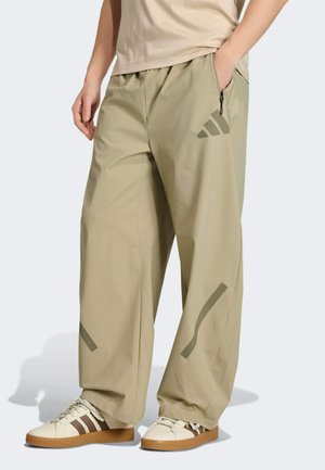 Pantaloni beige a gamba larga con vita a coulisse, dotati di una tasca con zip e dettagli scuri decorati. Indossati con sneakers beige e marroni.