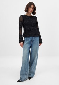 Zwarte lange mouwen top met bloemenkant details over doorschijnende stof, gecombineerd met wijde lichtblauwe denim jeans en puntige zwarte schoenen.