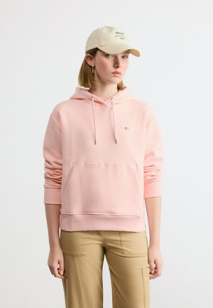 Jeune femme portant un sweat à capuche rose clair, un pantalon beige, une casquette beige et des boucles d'oreilles en or, debout devant un fond blanc uni.