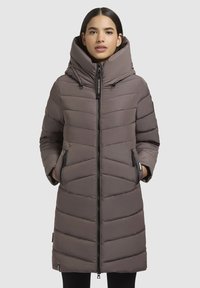 Femme portant un long manteau d'hiver matelassé marron avec un col montant et deux poches latérales zippées, se tenant devant un fond uni.