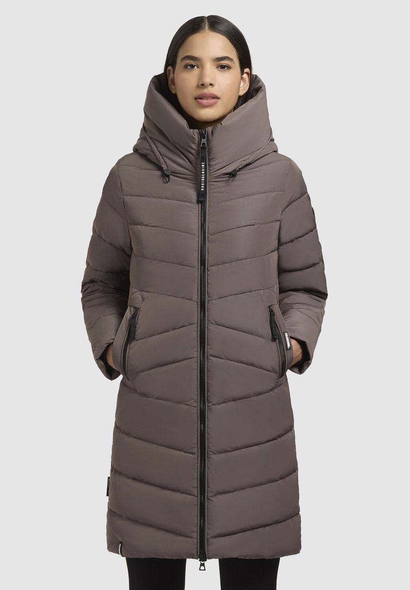 Femme portant un long manteau d'hiver matelassé marron avec un col montant et deux poches latérales zippées, se tenant devant un fond uni.