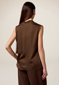 Blusa sin mangas de color marrón en material similar a la seda, con escote redondo y una abertura en la parte trasera, combinada con pantalones marrones a juego.