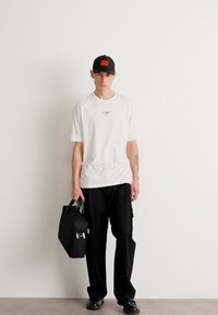 Hvid t-shirt med et lille grafisk print, sorte cargo-bukser, sort kasket med rød lap, holder en lille sort taske, sorte sko.