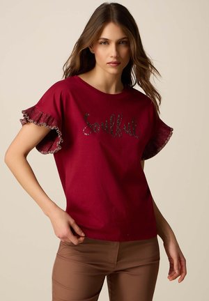 Ung kvinde med brunt hår iført en vinrød bluse med flæseærmer og teksten "Soulful", kombineret med lysebrune tætsiddende bukser mod en ensfarvet baggrund.