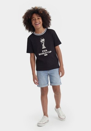 Smilende barn med krøllet hår iført sort FIFA World Cup t-shirt, lyse denimshorts og hvide sneakers, stående mod en ensfarvet baggrund.
