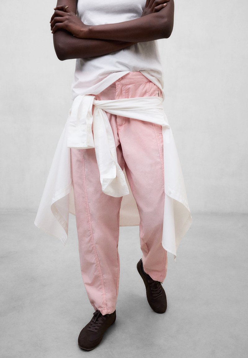 Personne les bras croisés portant un haut blanc sans manches, un pantalon rose, des chaussures noires et une chemise blanche nouée autour de la taille, debout sur un sol gris.