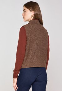 Gilet in maglia marrone con colletto alto, abbinato a una maglietta a maniche lunghe color ruggine. La texture è a costine e l'outfit è completato da jeans blu scuro.