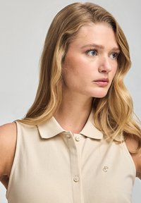 Beige ärmlös polo skjorta med krage, med en knappslå och broderad logotyp på vänster bröstområde. Slätt, lättviktigt tyg.