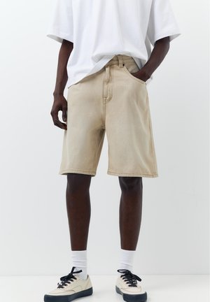 Személy bézs térdig érő rövidnadrágot, fehér, túlméretezett pólót, fehér zoknit és bézs tornacipőt fekete talppal és fűzőkkel visel, kezét a zsebében tartva áll.