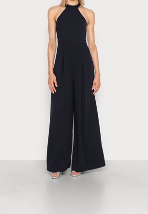 Navyblauwe jumpsuit met halternek, wijde benen en plooien. Heeft een gladde textuur en wordt gedragen met zilveren hoge hakken sandalen.