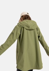 Light olive green waterproof jacket with a hood, featuring a simple design, smooth texture, and side pockets. Cap worn backward.