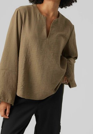 Personne portant un haut brun ample et texturé à manches longues avec un petit col en V, associé à un pantalon noir, sur un fond gris uni.