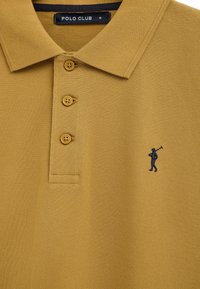 Polo color senape con tre bottoni, piccolo logo di un giocatore di polo blu navy sul petto e etichetta "POLO CLUB" taglia M all'interno del colletto.