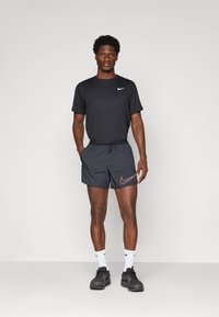Mann in schwarzem Nike-Laufoutfit mit Shorts, T-Shirt, weißen Socken und schwarzen Laufschuhen, der vor einem schlichten Hintergrund steht.