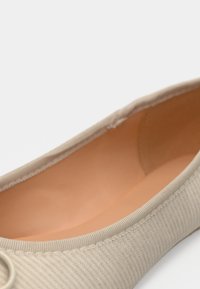 Ballerine en textile beige avec doublure intérieure lisse, présentant un extérieur côtelé et un design à bout arrondi. Accentuée par une décoration circulaire.