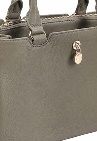 Sac à main en cuir gris avec une surface texturée, fermeture éclair et étiquette métallique décorative. Il possède des poignées doubles et des accents en métal doré.