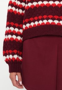 Knitted sweater with red, white, and burgundy zigzag patterns, featuring loose sleeves and a ribbed hem, paired with a maroon skirt.