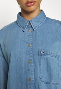 Camicia in denim azzurro chiaro con colletto a punta, bottoni a pressione argentati, tasca a toppa con dettagli logo e vestibilità rilassata e oversize.