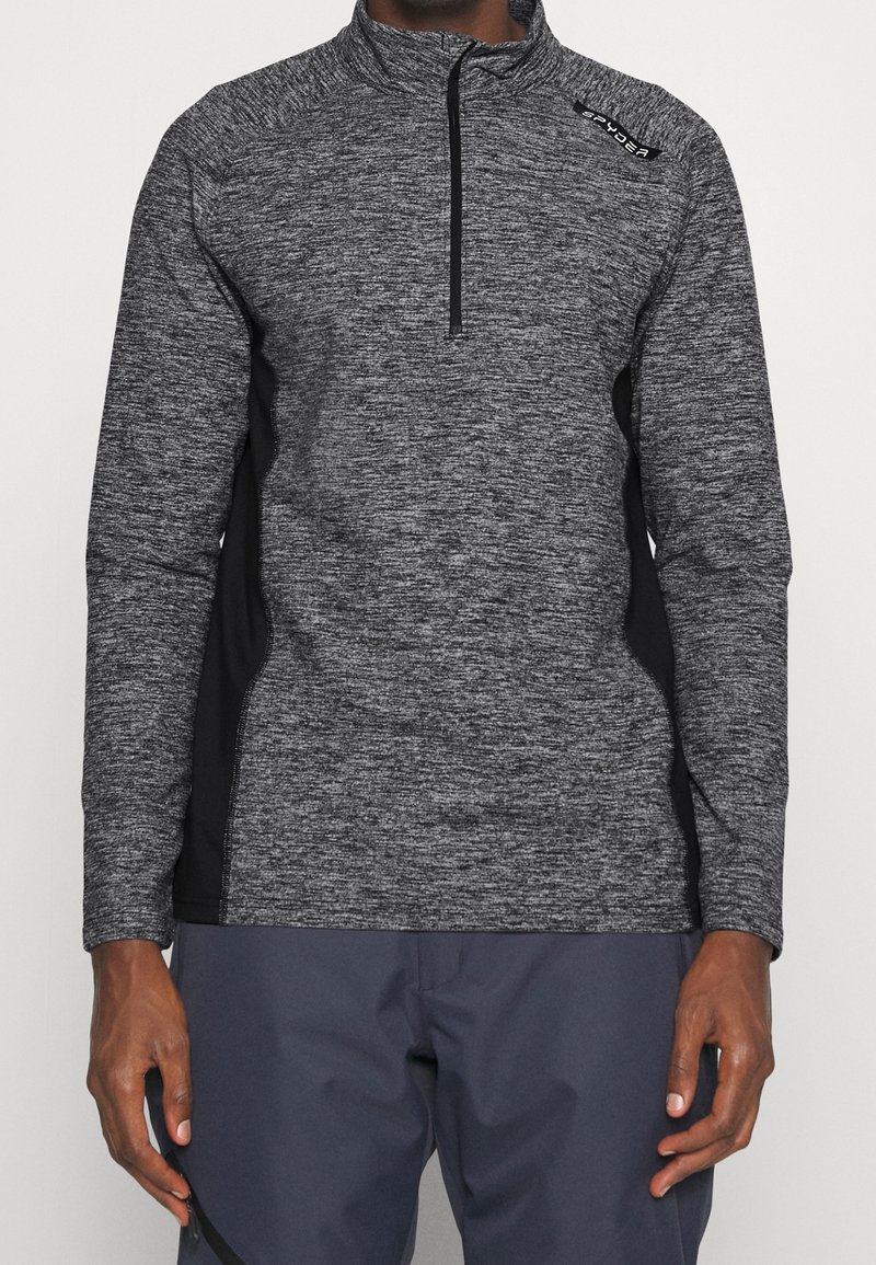 Pull-over à col zippé noir et gris en tissu tricoté texturé, avec des panneaux latéraux noirs et un petit logo sur l'épaule.