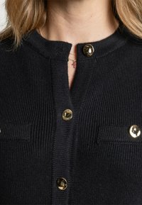 Schwarze gerippte Strickjacke mit rundem Ausschnitt, goldenen Knöpfen und zwei Fronttaschen. Die Textur ist weich mit einem gestrickten Muster.