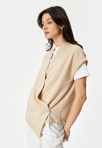 Gilet beige oversize con finitura testurizzata, caratterizzato da un davanti sovrapposto e una chiusura a un bottone su un lato, indossato sopra una camicia bianca.