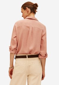 Una camicia rosa chiaro con maniche arrotolate, una yoke posteriore e una texture morbida; abbinata a pantaloni beige e una cintura marrone.